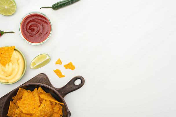 vrchní pohled na kukuřičné nachos s vápnem, chilli papričkami, kečupem a sýrovou omáčkou na dřevěné řezací desce na bílém pozadí - Fotografie, Obrázek