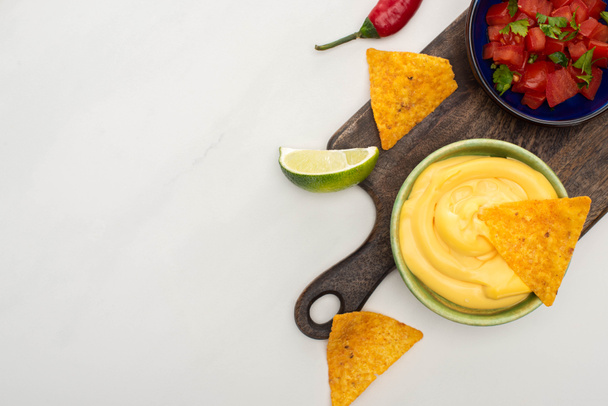 vrchní pohled na kukuřičné nachos s vápnem, chilli a sýrovou omáčkou na dřevěné řezací desce na bílém pozadí - Fotografie, Obrázek