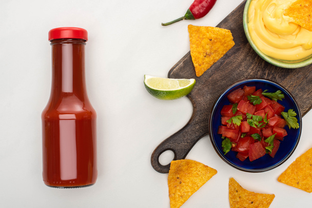 top view of corn nachos with lime, chili, ketchup and cheese sauce on wooden cutting board on white background - Foto, afbeelding
