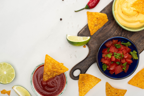 top view of corn nachos with lime, chili, ketchup and cheese sauce on wooden cutting board on white background - Photo, Image