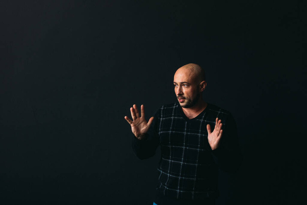 a young man with glasses, in dark clothes, bald, on a black background, shows something with his hands with emotions on his face - 写真・画像