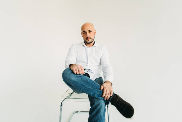 a young man in glasses, in a white shirt and a dandy, bald, sits on a chair on a white background, shows something with his hands with emotions on his face - Foto, immagini