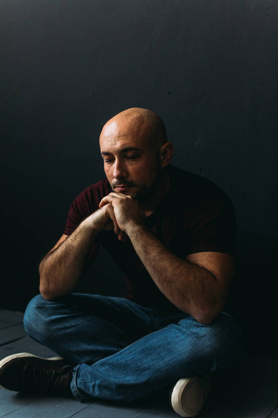 a young man with glasses, in jeans and a T-shirt, bald, sitting on the floor against a black wall - Photo, image