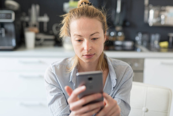 Worried woman indoors at home kitchen using social media apps on phone for video chatting and stying connected with her loved ones. Stay at home, social distancing lifestyle - Φωτογραφία, εικόνα