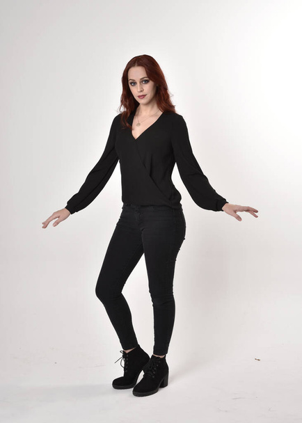 Portret van een mooi meisje met rood haar in zwarte jeans, laarzen en een blouse. full length staande houding, met uitzicht op de camera met handgebaren op een studio achtergrond. - Foto, afbeelding