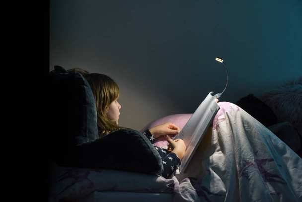 Κορίτσι που διαβάζει ένα βιβλίο στο κρεβάτι το βράδυ στο άνετο υπνοδωμάτιο φορώντας πιτζάμες - Φωτογραφία, εικόνα