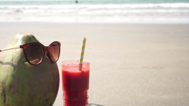 Coconut in sunglasses at the sea with a glass of juice. Tropical beach - Footage, Video