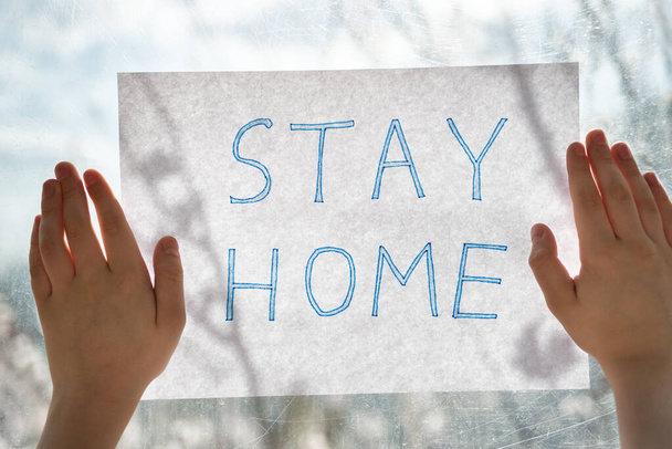Mãos de crianças segurando folha de papel com inscrição STAY HOME na janela. Conceito de isolamento de quarentena
. - Foto, Imagem