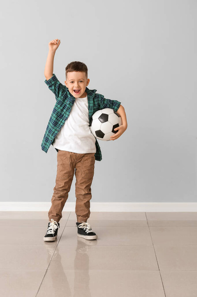Niño feliz con pelota de fútbol cerca de la pared gris
 - Foto, Imagen