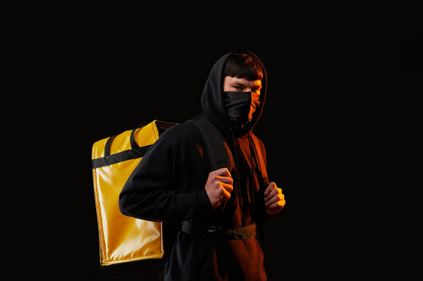 オレンジ色の光で黒の背景に黄色のバックパックを持つゴムマスクの白い服の若い配達人 - 写真・画像
