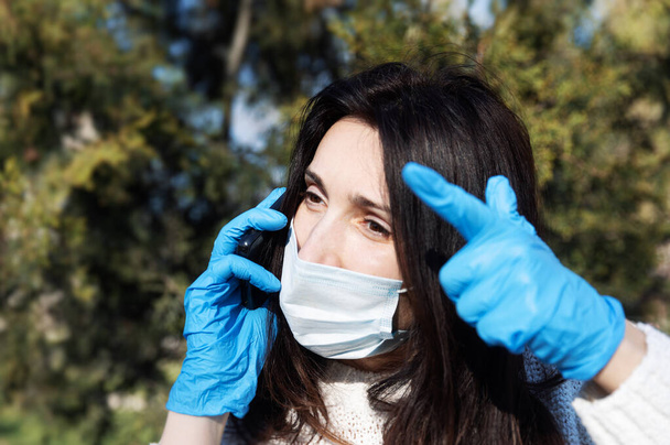 Jovem mulher em uma máscara médica com um telefone celular se comunica emocionalmente e gestos
 - Foto, Imagem