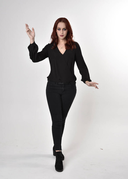 Porträt eines hübschen Mädchens mit roten Haaren, das schwarze Jeans, Stiefel und eine Bluse trägt. in voller Länge stehend, mit Handgesten auf Studiohintergrund in die Kamera gerichtet. - Foto, Bild