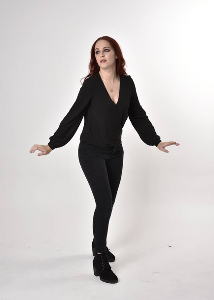 Portrait d'une jolie fille aux cheveux roux portant un jean noir, des bottes et un chemisier. pose debout sur toute la longueur, face à la caméra avec des gestes de main sur un fond de studio
. - Photo, image
