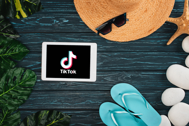KYIV, UKRAINE - MARCH 25, 2020: Top view of digital tablet with TikTok app near flips flops, straw hat with pebbles and leaves on dark wooden surface - Zdjęcie, obraz