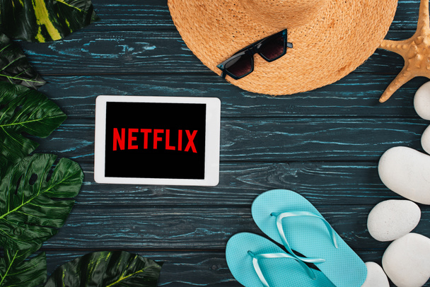 KYIV, UKRAINE - MARCH 25, 2020: Top view of digital tablet with netflix app near straw hat, flip flops and leaves on dark wooden surface - Foto, immagini