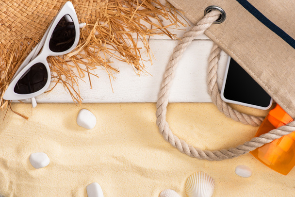 Top view of smartphone and sunscreen in bag near straw hat and sunglasses on white wooden planks and pebbles on sand - 写真・画像