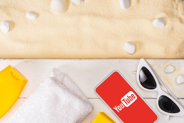 KYIV, UKRAINE - MARCH 25, 2020: Top view of smartphone with youtube app near sunglasses, sunscreen and towel on white wooden planks and pebbles on sand  - 写真・画像