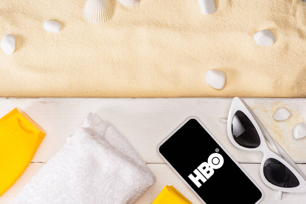 KYIV, UKRAINE - MARCH 25, 2020: Top view of smartphone with hbo app near sunglasses, sunscreen and towel on white wooden planks near pebbles on sand  - Фото, изображение