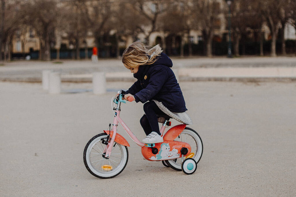 13:02:2020 Paris, France: little girl with blond hair rides quickly on a pink bike without helmet and fall protection down the street in cloudy autumn weather - Photo, image