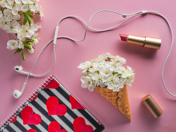 white cherry flowers into waffle cone with accessories and cosmetic lipstick and scetchbook with speakerphones pink background - Фото, изображение