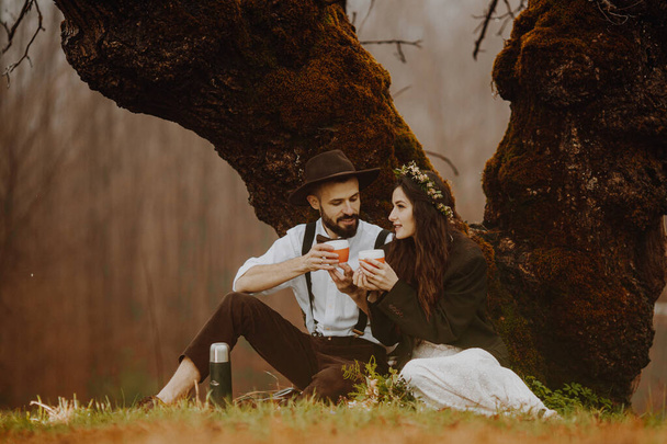 young loving couple outdoors drinking hot coffee or tea hugging while sitting under a tree. copy space - Fotoğraf, Görsel