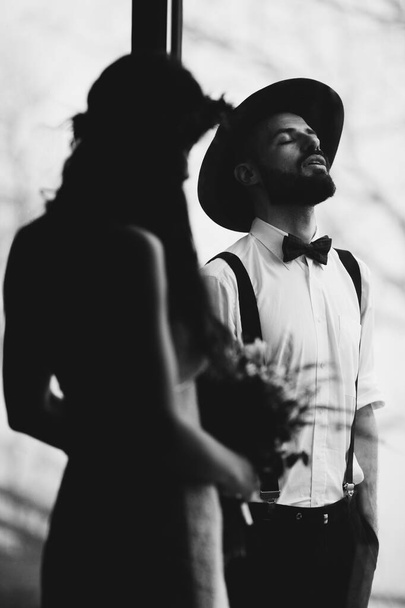 portrait of a stylish wedding couple: Young bearded man in hat and brunette bride in elegant gown wearing a wreath braided in hairstyle - Photo, image