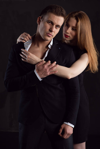 Sexy red-haired girl unzips the shirt of a brutal man in a black suit in a fit of passion - Фото, изображение