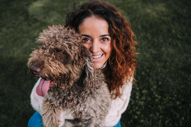 A beautiful woman is in the meadow with her dog. The owner is hugging her pet while she is looking at the camera. They are enjoying a day in the park. The pet is a Spanish water dog with brown fur. - Фото, изображение