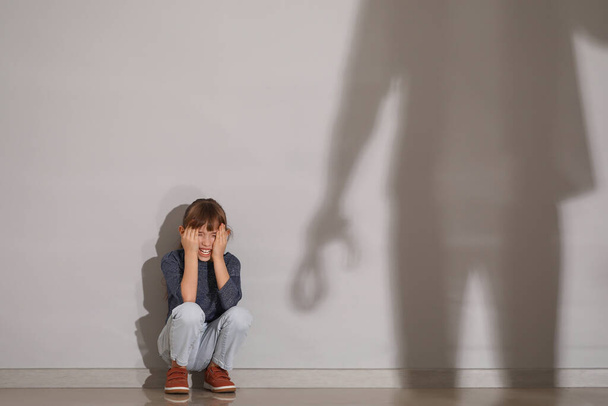 Σκιά του πατέρα απειλεί την κορούλα του που κάθεται στο πάτωμα κοντά στον γκρίζο τοίχο - Φωτογραφία, εικόνα