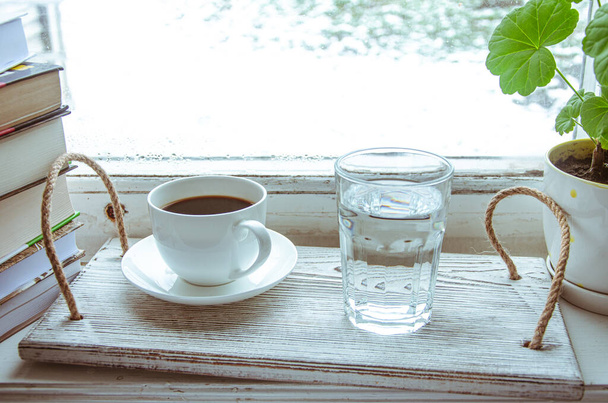 A white cup of coffee and a glass of water stand on a wooden tray on the windowsill next to a stack of books. Close-up - Foto, Imagen