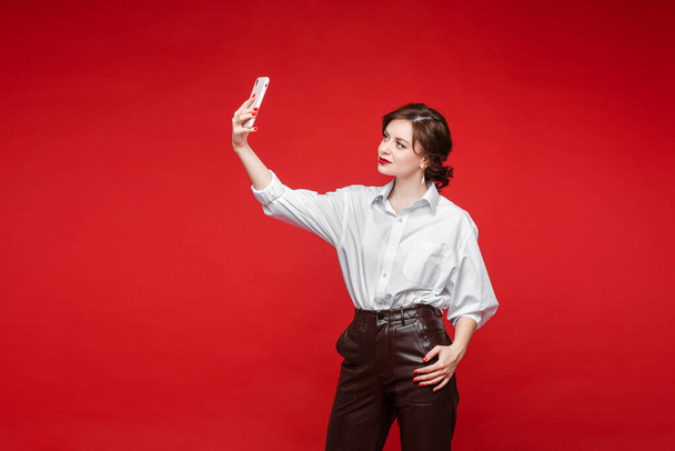 Handsome female holds a phone and takes a selfie, picture isolated on red background - Fotografie, Obrázek