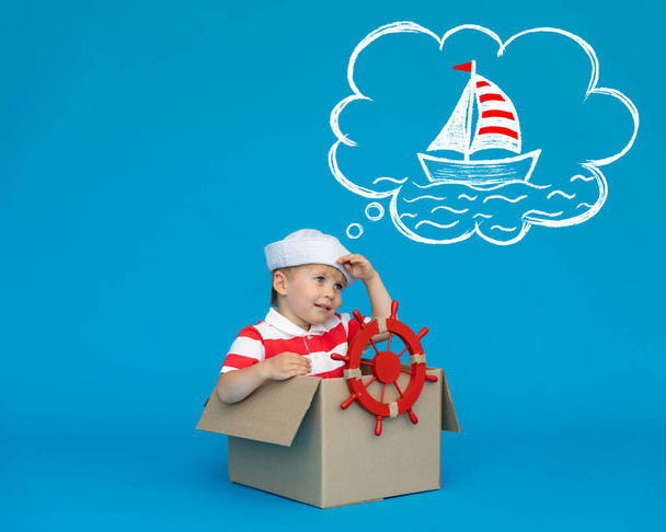 Happy child dreams of becoming a captain. Kid having fun against blue background. Boy wearing striped shirt playing in cardboard box. Summer vacation and travel concept. Dream and imagination - Foto, imagen