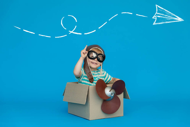 Happy child dreams of becoming a pilot. Kid having fun against blue paper background. Boy wearing striped shirt playing in cardboard box. Summer vacation and travel concept. Dream and imagination - 写真・画像