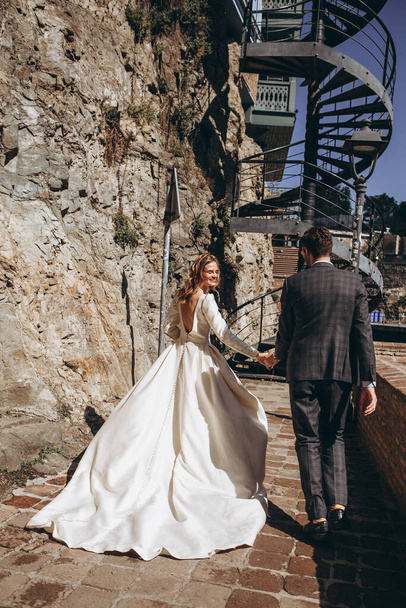 01.06.2019 Tbilisi, Georgia: Georgian-style wedding: Stylish mustachioed man and attractive blonde bride in pretty dress posing for photo on old city streets - Photo, Image