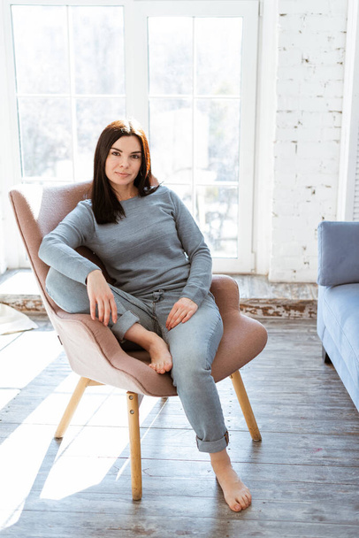 portrait of a young woman in jeans clothes sitting in a pink armchair in front of a window at home - Foto, Imagen