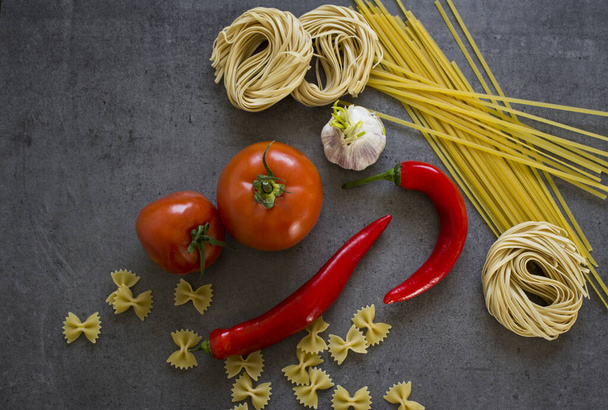 Beautiful still life with pasta of different types and fresh vegetables on stone table. Italian food ingredients on dark grey background. Regional food close up photo. Vibrant colors. - Photo, Image