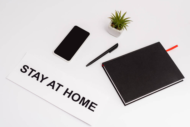 smartphone with blank screen near pen, notebook, plant and paper with stay at home lettering on white  - Foto, immagini