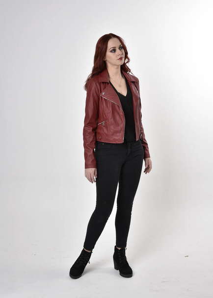 Portrait of a pretty girl with red hair wearing black jeans and boots with leather jacket.  full length standing pose on a studio background. - Photo, Image