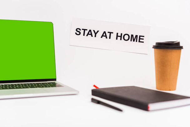 selective focus of laptop with green screen near paper cup, notebook, pen and paper with stay at home lettering isolated on white  - Photo, Image
