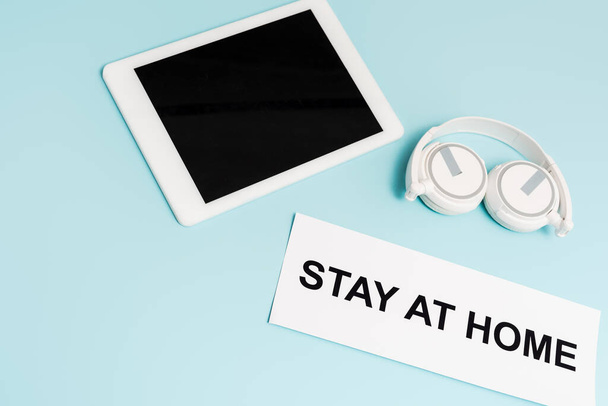 digital tablet with blank screen near headphones and paper with stay at home lettering on blue  - Photo, Image