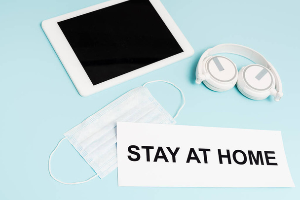 tablet digital com tela em branco perto de fones de ouvido, máscara médica e papel com estadia em casa lettering em azul
  - Foto, Imagem