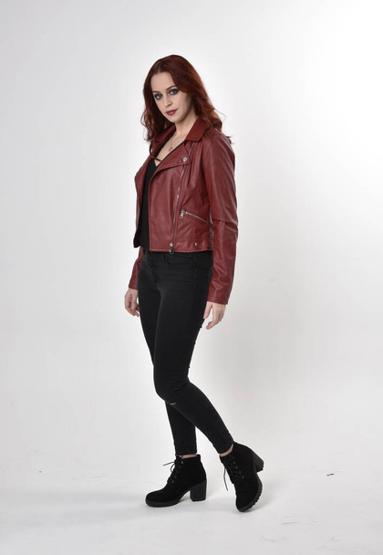 Portrait of a pretty girl with red hair wearing black jeans and boots with leather jacket.  full length standing pose on a studio background. - Photo, image