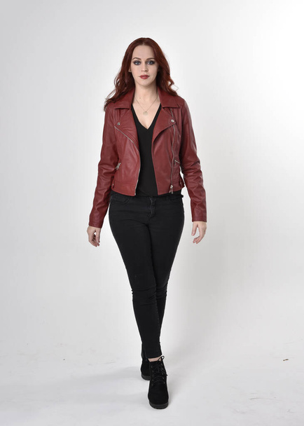 Porträt eines hübschen Mädchens mit roten Haaren, das schwarze Jeans und Stiefel mit Lederjacke trägt. Stehende Pose in voller Länge auf einem Studiohintergrund. - Foto, Bild