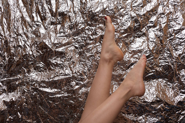 Beautiful female feet on a beautiful background in the form of a foil. Fashionable photo of female legs. Red pedicure. - Photo, Image