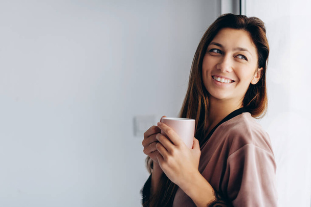 Usmívající se dívka u okna. Bruneta pije svou ranní kávu. Zůstat doma, útulné ráno. - Fotografie, Obrázek