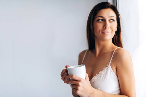 Smiling girl by the window. Brunette girl is drinking her morning coffee. Staying home,cozy morning. - Photo, Image