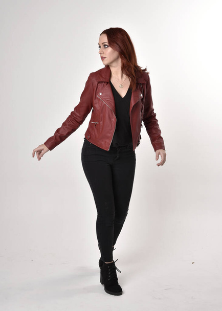 Portrait d'une jolie fille aux cheveux roux portant un jean noir et des bottes avec une veste en cuir. pose debout pleine longueur avec geste de la main sur un fond de studio
. - Photo, image
