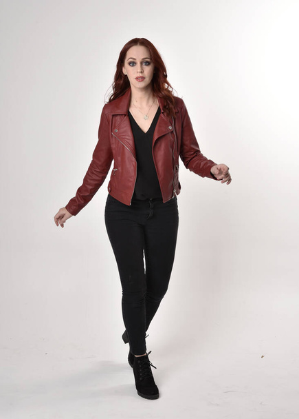 黒のジーンズと革のジャケットとブーツを身に着けている赤い髪のきれいな女の子の肖像画。スタジオの背景に手のジェスチャー付きの全長立ちポーズ. - 写真・画像