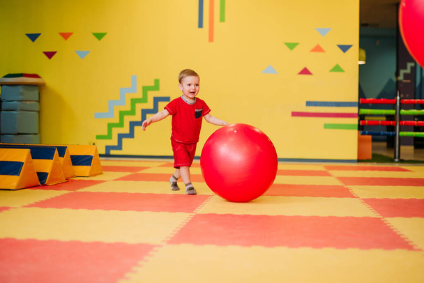 Happy laughing boy 1-2 years old having fun in ball pit on birthday party in kids amusement park and indoor play center. Child playing with colorful balls in playground ball pool. Activity toys for little kid. - Photo, Image