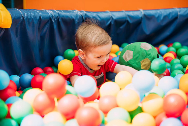 Happy laughing boy 1-2 years old having fun in ball pit on birthday party in kids amusement park and indoor play center. Child playing with colorful balls in playground ball pool. Activity toys for little kid. - Foto, Imagem